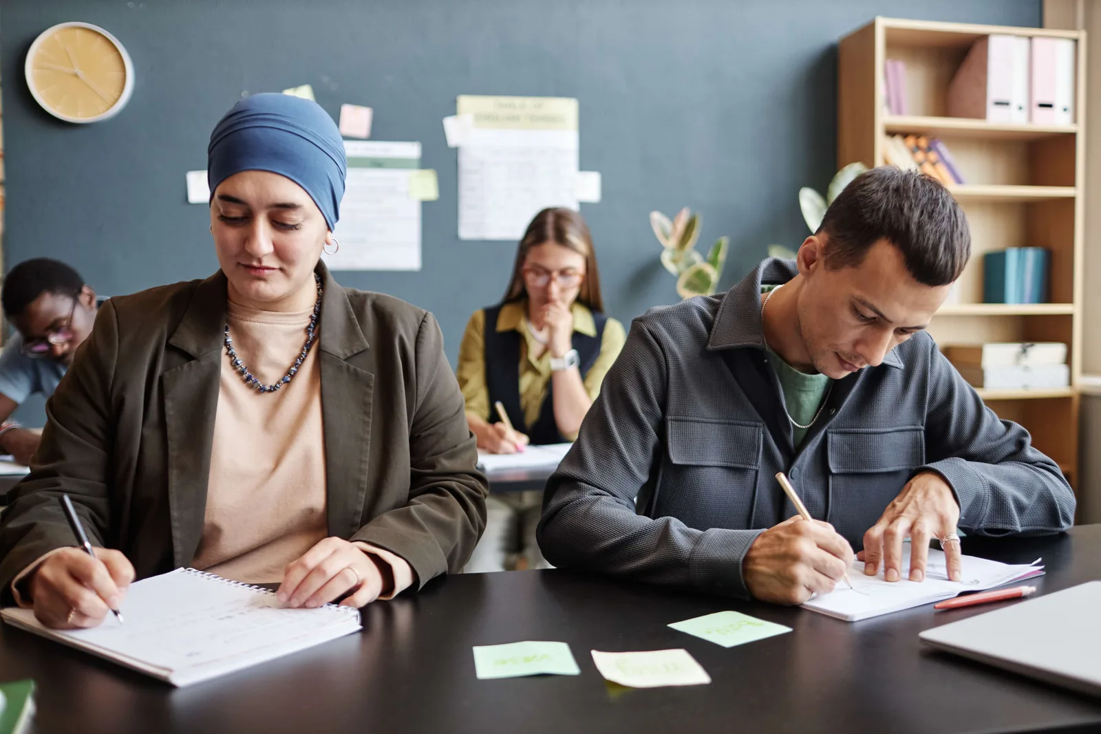 Muslim Woman Writing in Notebook Sitting at desk next to Man writing i n classroom