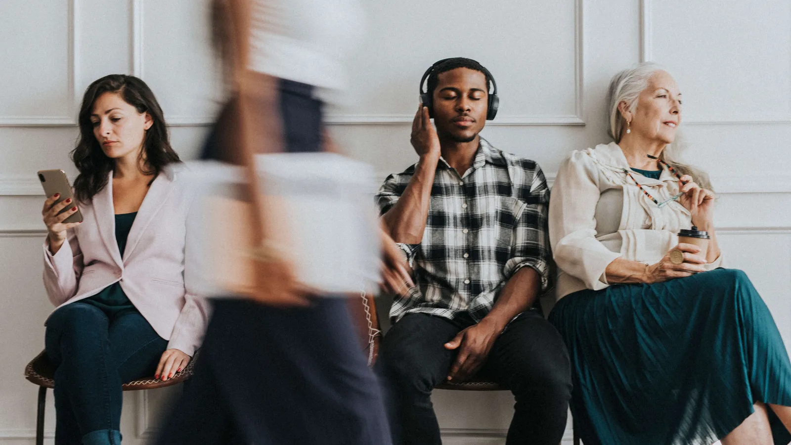 Diverse people sitting by a white wall on phones and listening to different devices