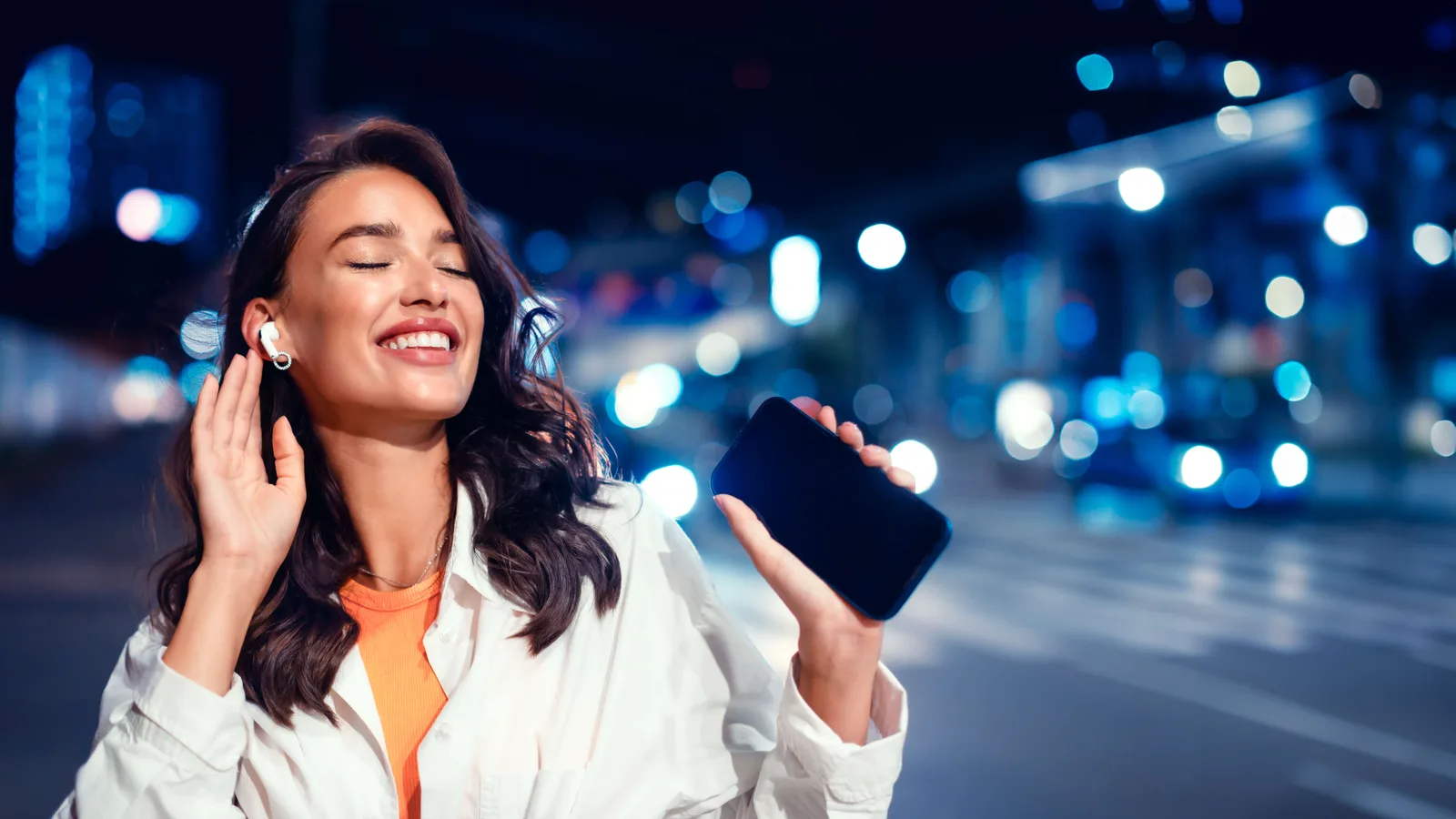 Joyful lady enjoying favourite music on radio in earphones holding smartphone, walking at night on city street