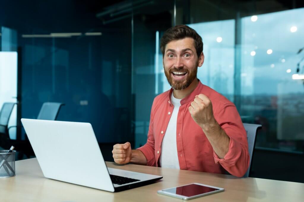 Portrait of successful mature businessman in red shirt at workplace inside office, man celebrating victory and successful achievement results at work, looking at camera, holding hand up.